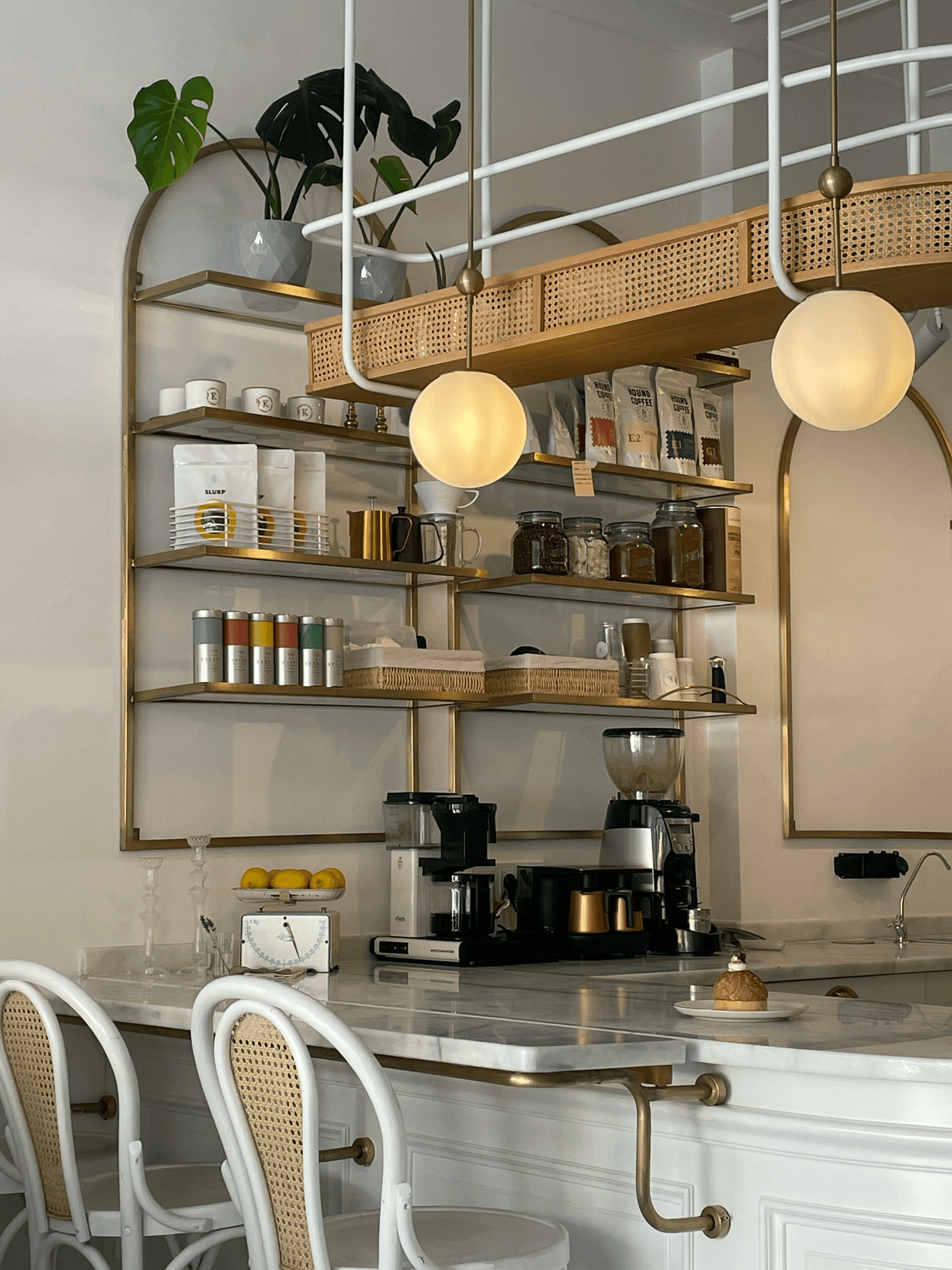 Picture of a coffee shop, with white marble on top of the bar. A display of various ingredients to makek beverages. Gold decorations on a white wall.