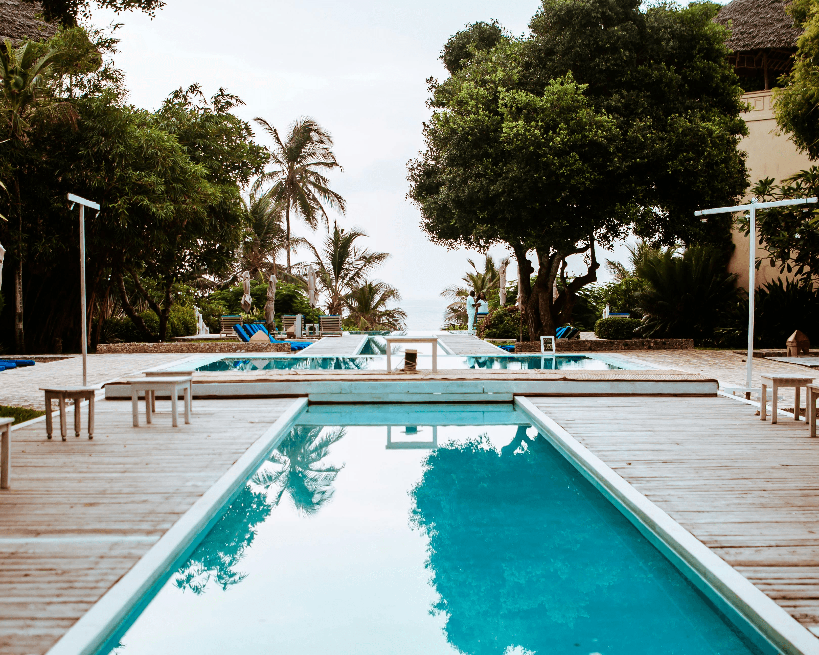 Picture of a pool in a hotel, surrounded by a spruce deck. Various trees behind the deck and sea in the far background