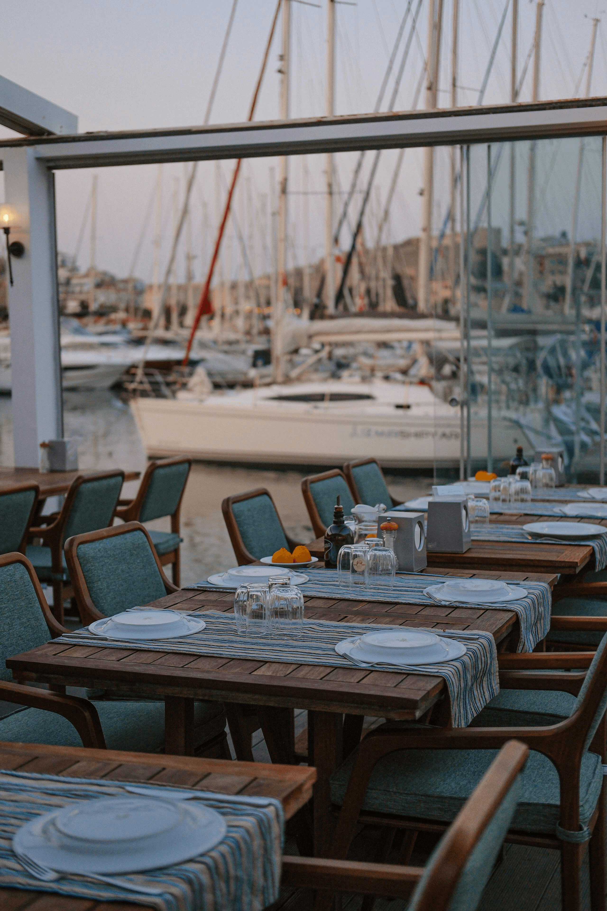 Picture of a nice restaurant next to a marina. Yachts on the background. The restaurant has wooden chairs with blue cloth on the cushions, and wooden tables with light blue decorations.